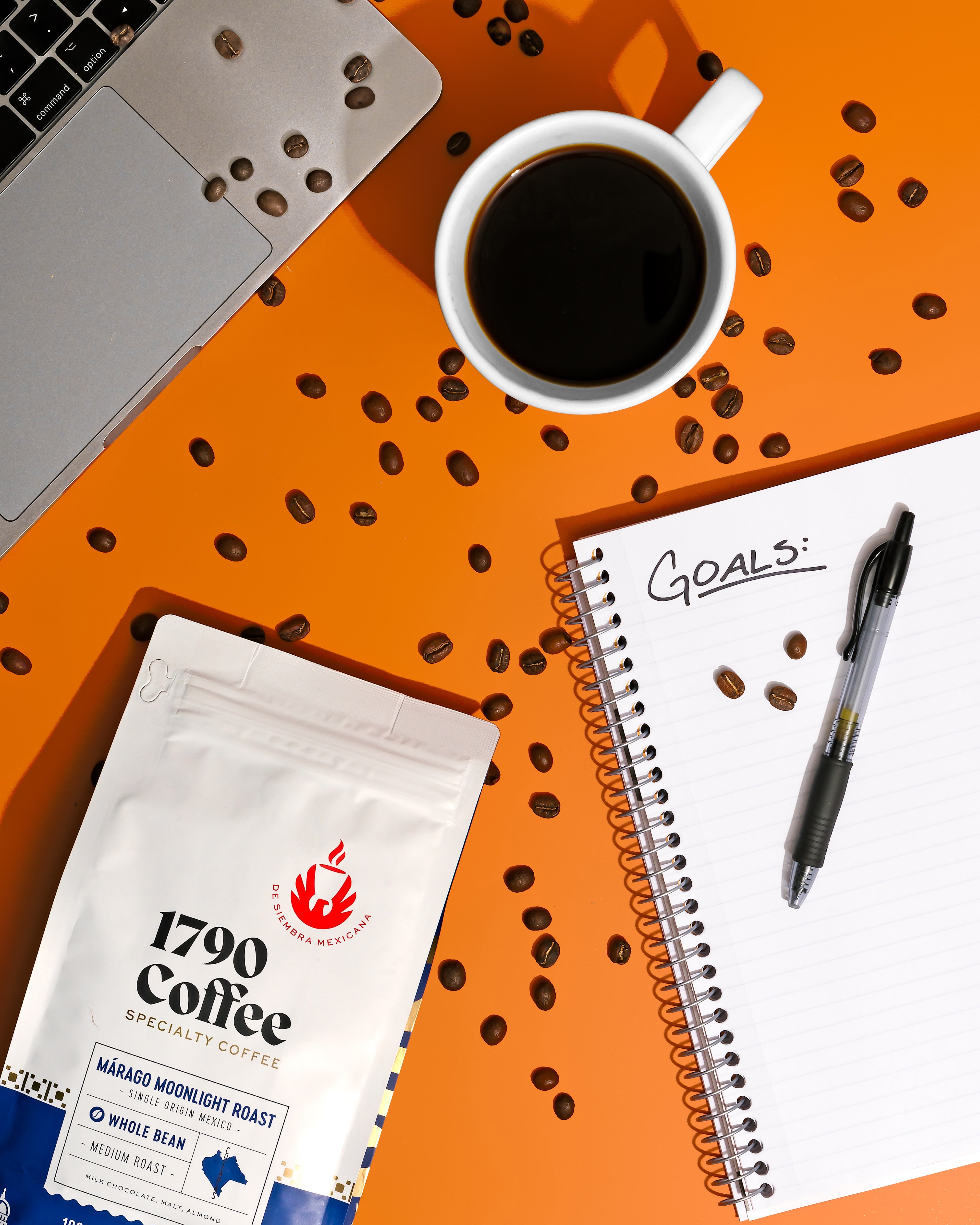 Bag of 1790 Coffee Marago Moonlight Roast with a cup of coffee, notebook, and pen on an orange surface with scattered coffee beans.
