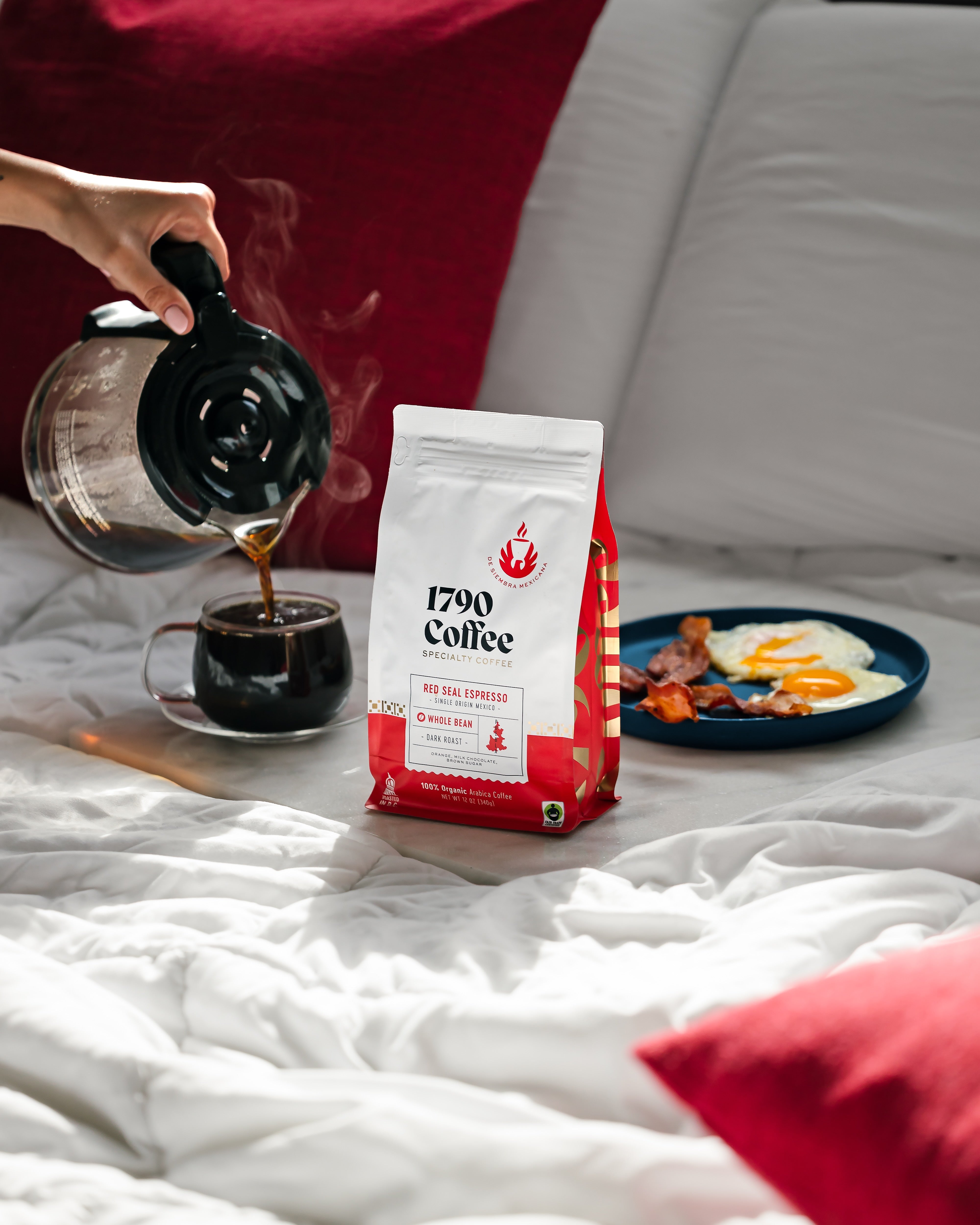 Person pouring coffee from a pot next to a bag of 1790 Coffee Red Seal Espresso on a bed with a plate of bacon and eggs.